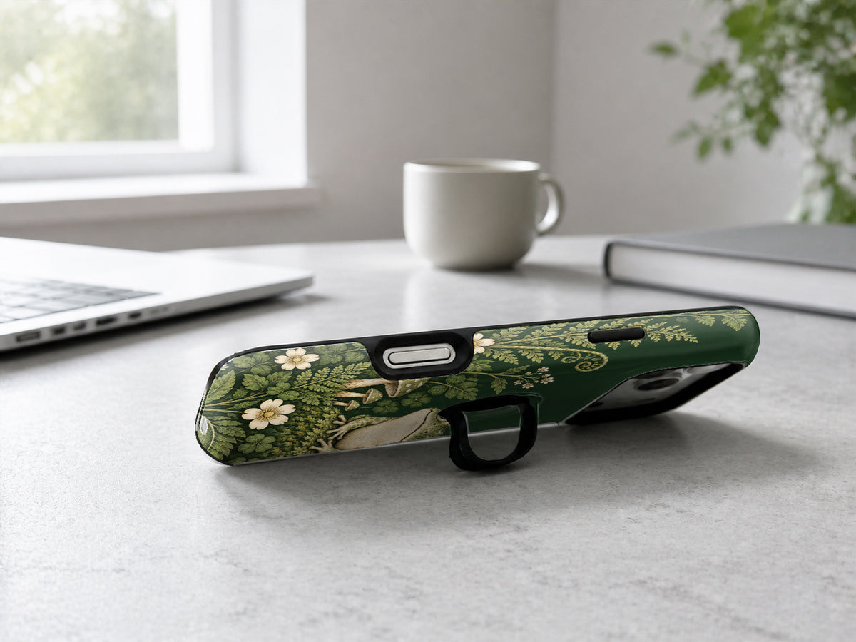 Forest Green Frog and Mushroom Garden MagSafe Loop iPhone Case, shown as a kickstand on a desk