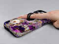 Purple Pressed Flower Garden MagSafe Loop iPhone Case shown in use with a finger through the horizontal loop